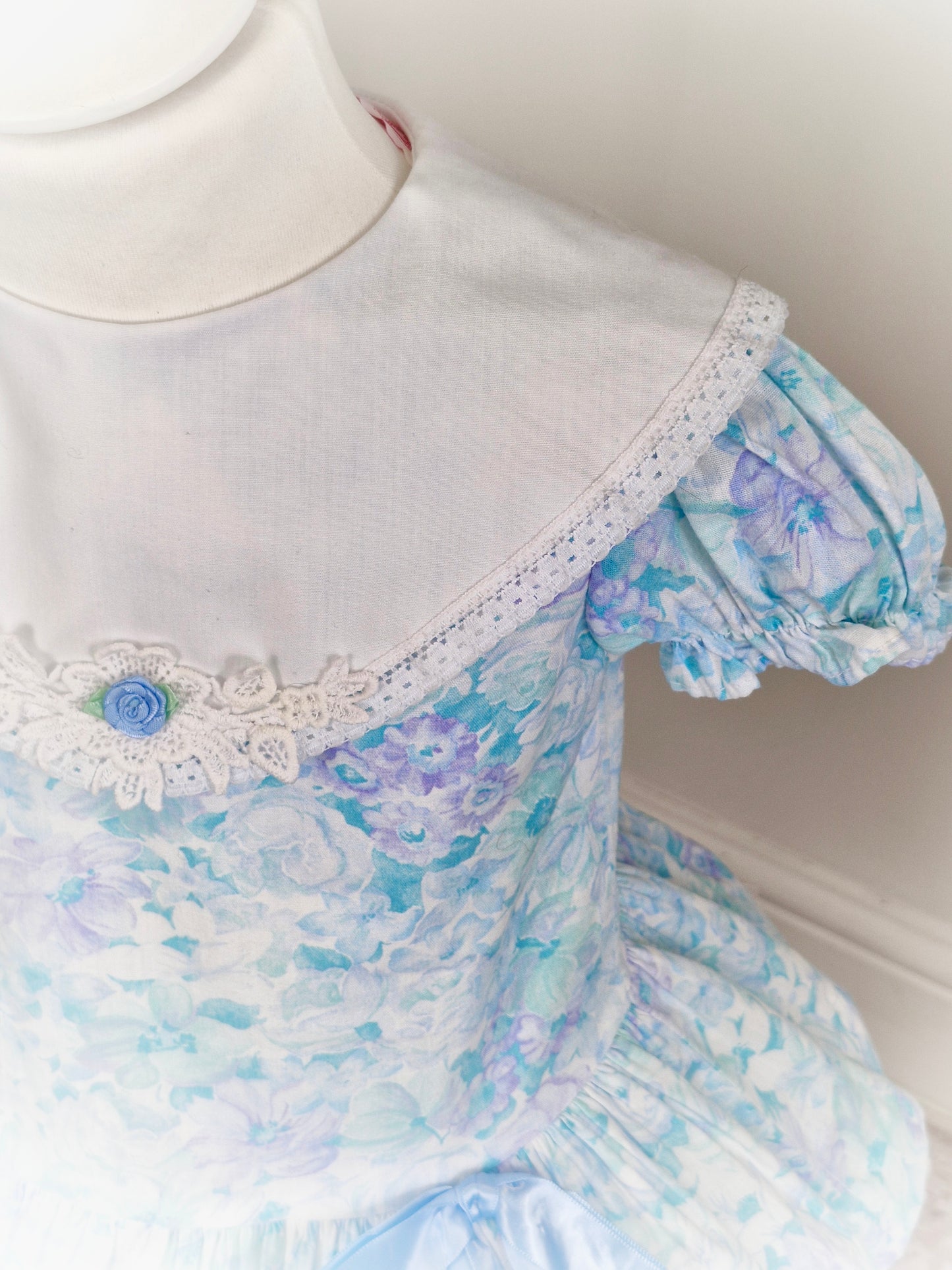 2 years. Pastel coloured drop waist Roanna with white bib and blue ribbon