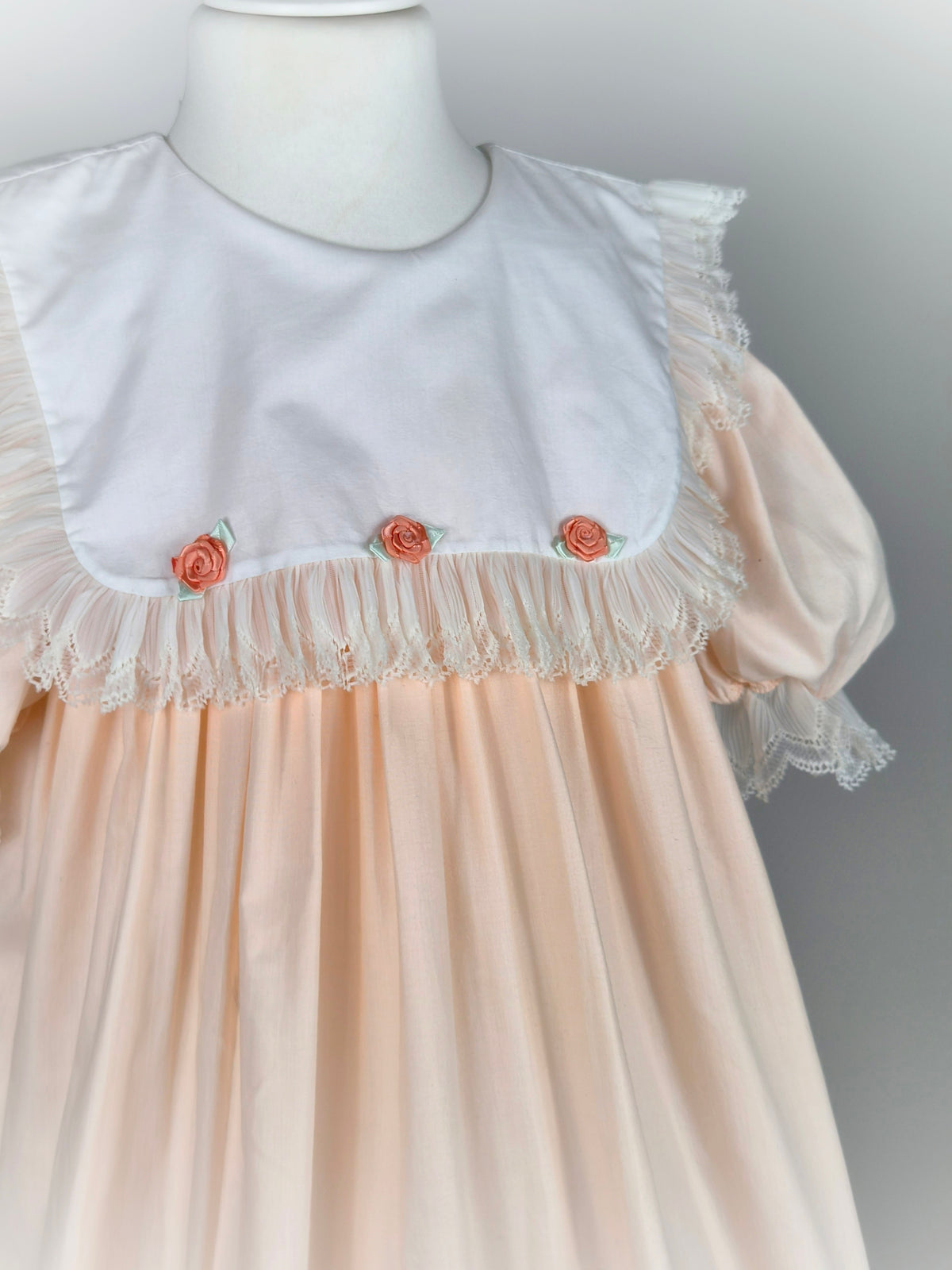 8-9 years. Peach vintage dress with white bib, lace trim and 3 peach coloured rosettes
