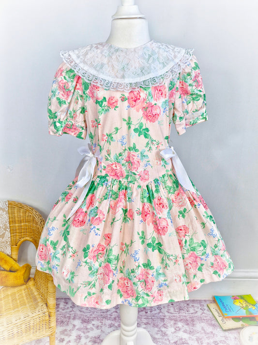 6-7 years. Peach dress with pink flowers, white lace bib and ribbon bows on sides