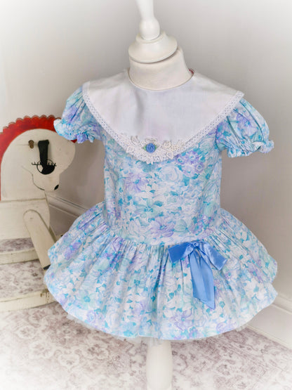 2 years. Pastel coloured drop waist Roanna with white bib and blue ribbon