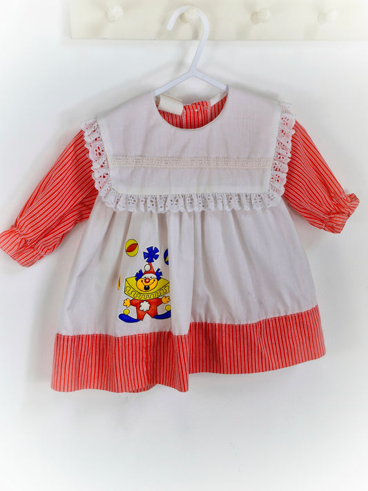 12 months. White dress with red sleeves and hem, lace trim on bib and clown applique