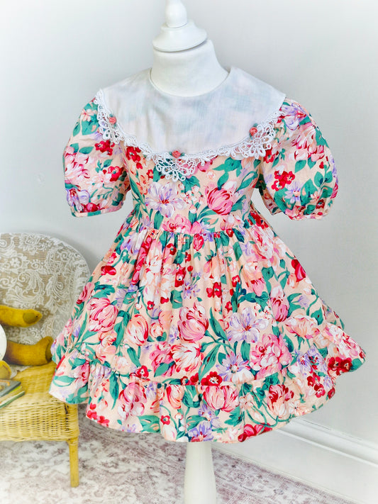 3 years. Peach vintage dress with large flower print, white bib with lace trim and 3 rosettes. Bonnie Jean.