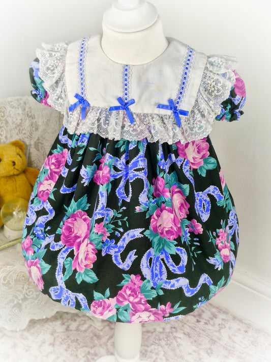 2 years. Black vintage Jolene dress with floral and ribbon print, white lace bib and blue bows