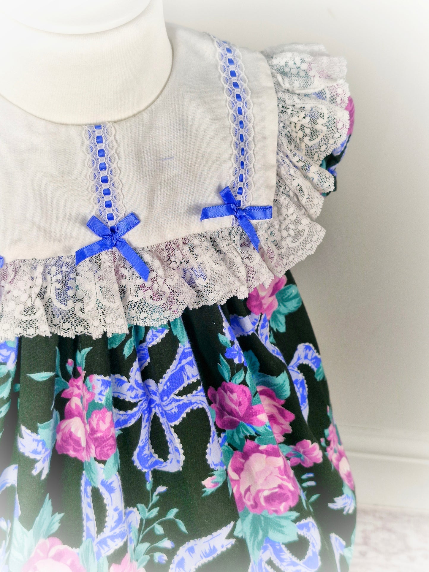 2 years. Black vintage Jolene dress with floral and ribbon print, white lace bib and blue bows