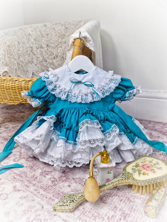 6-12 months. Teal Lid'l Dolly with white ruffles, lace and bib