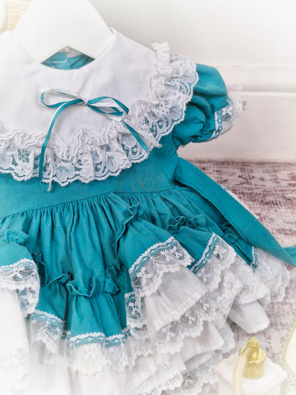 6-12 months. Teal Lid'l Dolly with white ruffles, lace and bib