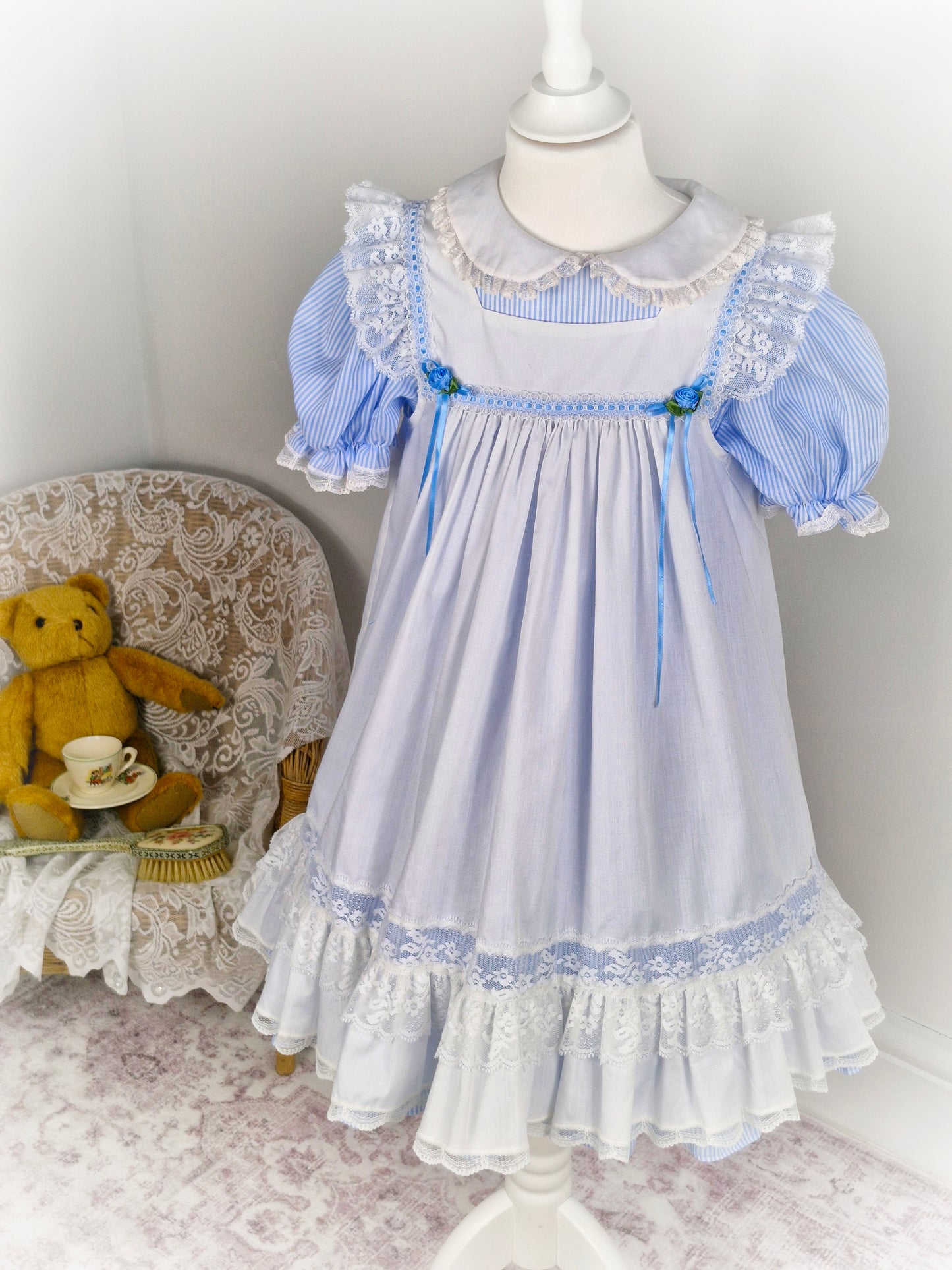 6-7 years. Blue and white candy striped dress with puff sleeves & white apron with flutter shoulders, lace hem and blue ribbons and rosettes