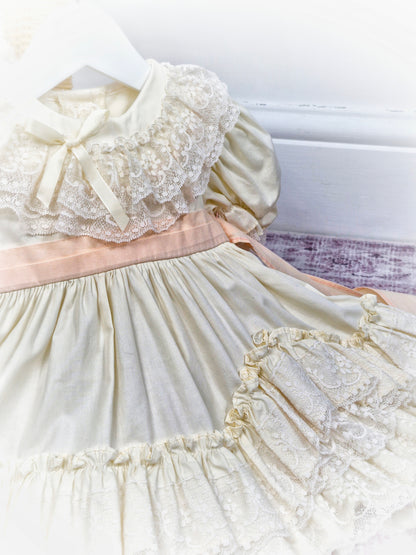 9-12 months. Cream vintage dress with lace, frills and layers and a peach tieback