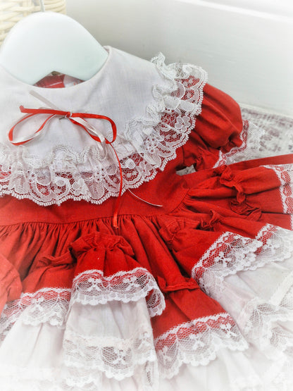 6-12 months. Red Lid'l Dolly with white bib and lace ruffles