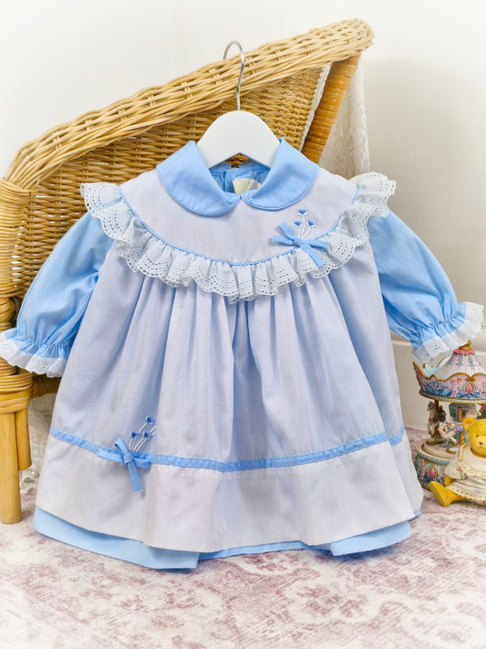 12-18 months. Two piece, blue dress with puff sleeves and Peter Pan collar with white apron, trimmed with white lace and blue embroidered flowers