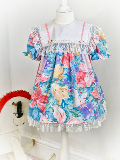 3-4 years. Watercolour printed vintage dress from Sandys Girls with large white bib, pink satin ribbon and rosettes and large lace trim