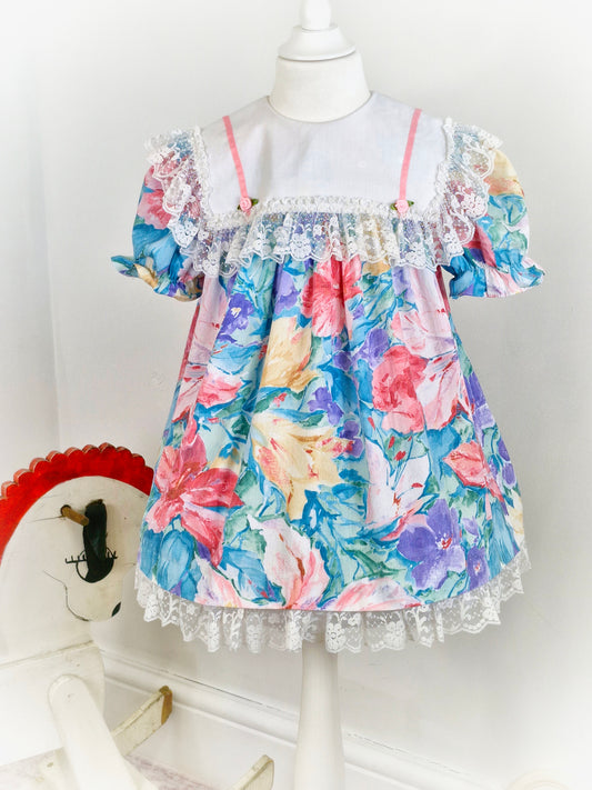 3-4 years. Watercolour printed vintage dress from Sandys Girls with large white bib, pink satin ribbon and rosettes and large lace trim