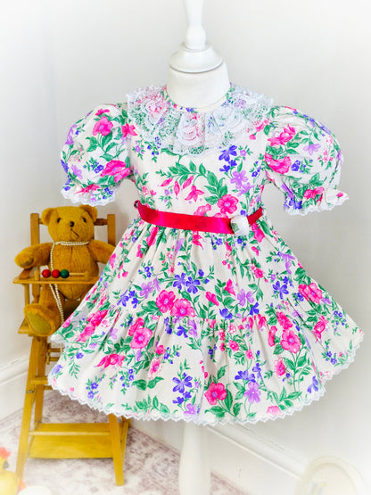 3 years. White vintage dress with vibrant floral print in pink, purple and green. White lace collar and hot pink satin waist tie