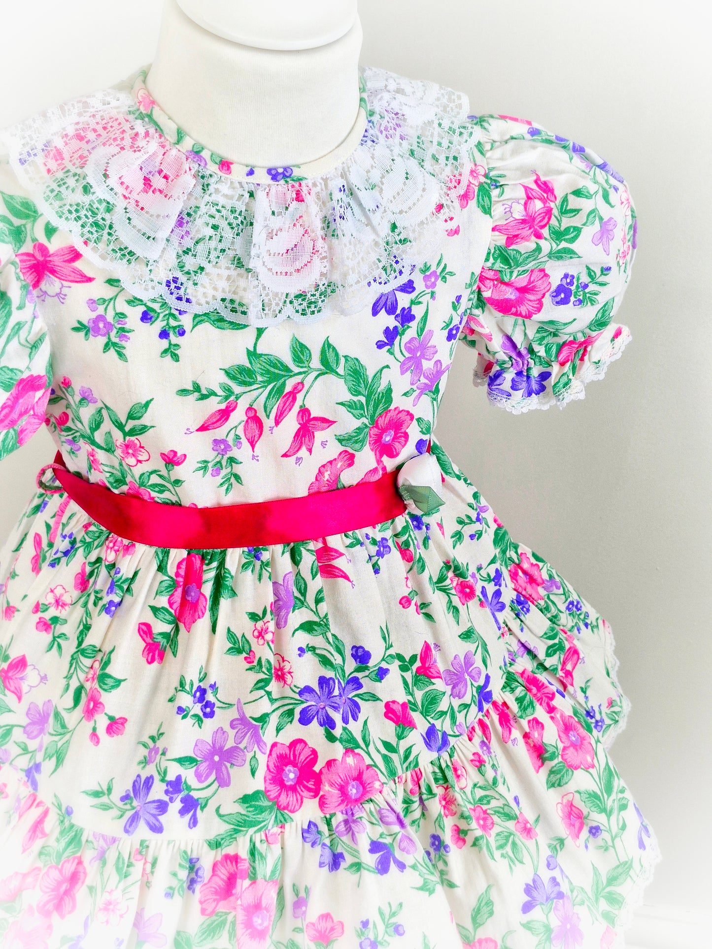 3 years. White vintage dress with vibrant floral print in pink, purple and green. White lace collar and hot pink satin waist tie