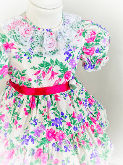 3 years. White vintage dress with vibrant floral print in pink, purple and green. White lace collar and hot pink satin waist tie