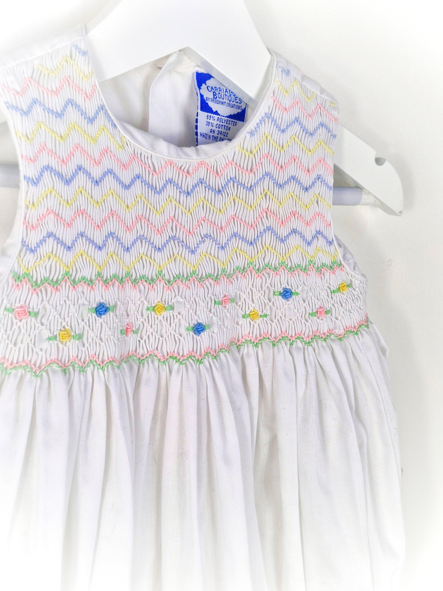 6-9 months. White sleeveless romper with smocking embroidered with pink, blue and lemon zigzags and flowers. Carriage Boutique