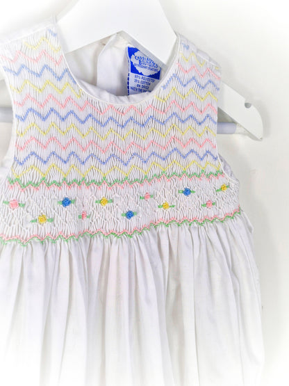 6-9 months. White sleeveless romper with smocking embroidered with pink, blue and lemon zigzags and flowers. Carriage Boutique