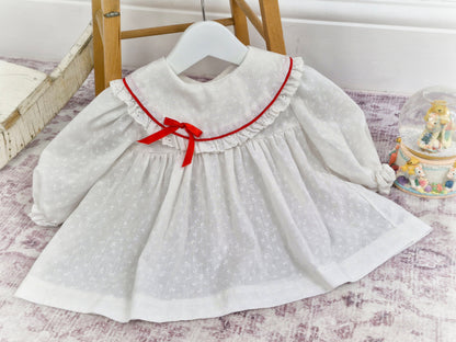 6-9 months. White vintage dress with long sleeves, white floral print, red ribbon and eyelet trimmed bib