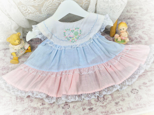 6-9 months. Blue and pink tiered sleeveless vintage dress with white bib, embroidered with floral heart and trimmed with lace