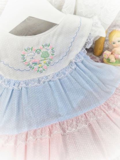 6-9 months. Blue and pink tiered sleeveless vintage dress with white bib, embroidered with floral heart and trimmed with lace