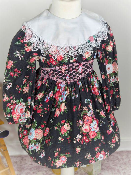 2-3 years. Black floral Polly Flinders smock dress with pink embroidery, white bib with lace trim and pink rosette