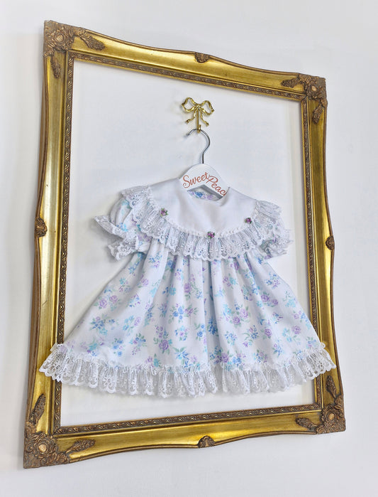 12 months. White Cutest One dress with blue and lilac flowers, lace trim and rosettes on bib