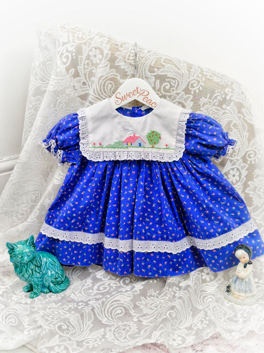 9-12 months. Blue dress with floral print and white bib embroidered with a home scene