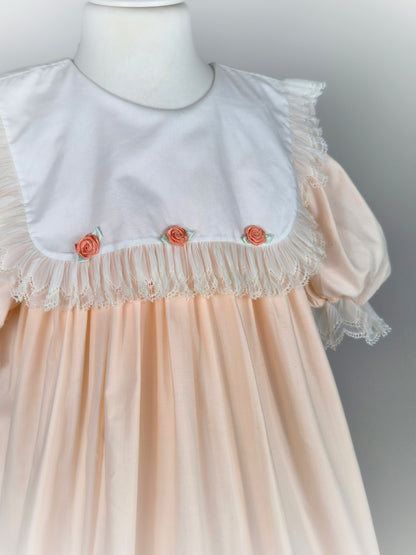 8-9 years. Peach vintage dress with white bib, lace trim and 3 peach coloured rosettes